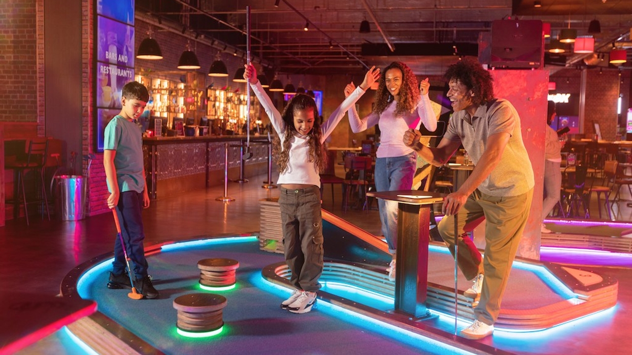 Family enjoying mini-golf indoors with neon lights and a bar in the background.