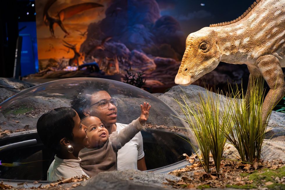 Family in a dome views a dinosaur exhibit with a prehistoric backdrop.