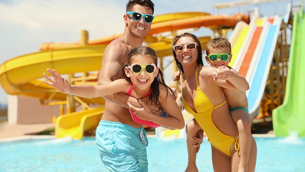 Family in swimsuits and sunglasses enjoying a water park with colorful slides behind them.