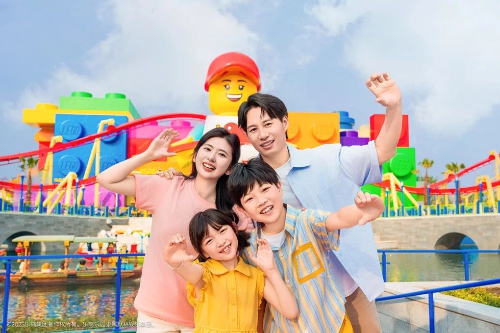 Family of four waving at colorful theme park with a giant LEGO figure.