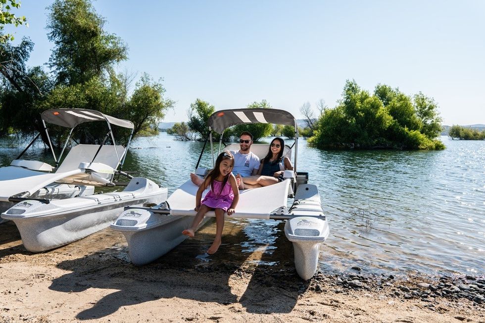 Family on a motorised chaise lounge by a lake shore, with lush greenery and clear blue sky in the background.