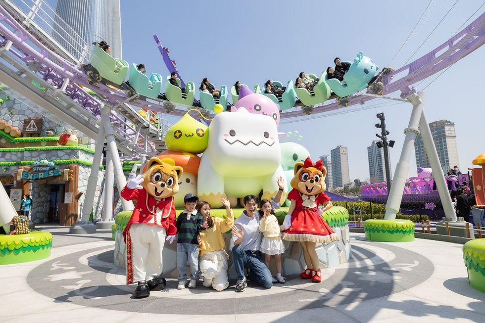 Family posing with mascots at a colorful amusement park ride under a clear sky.