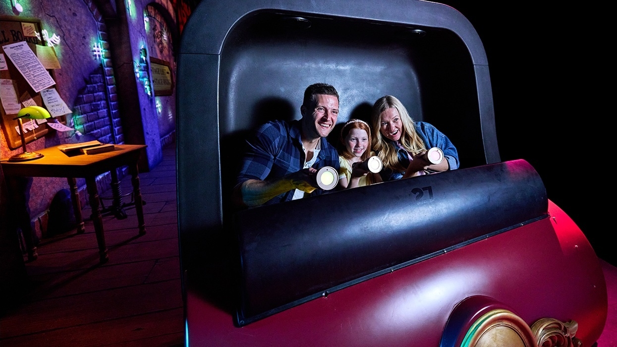 Family smiling on a themed amusement ride called 'Phantom Theater: Opening Nightmare' with flashlights, colorful background.