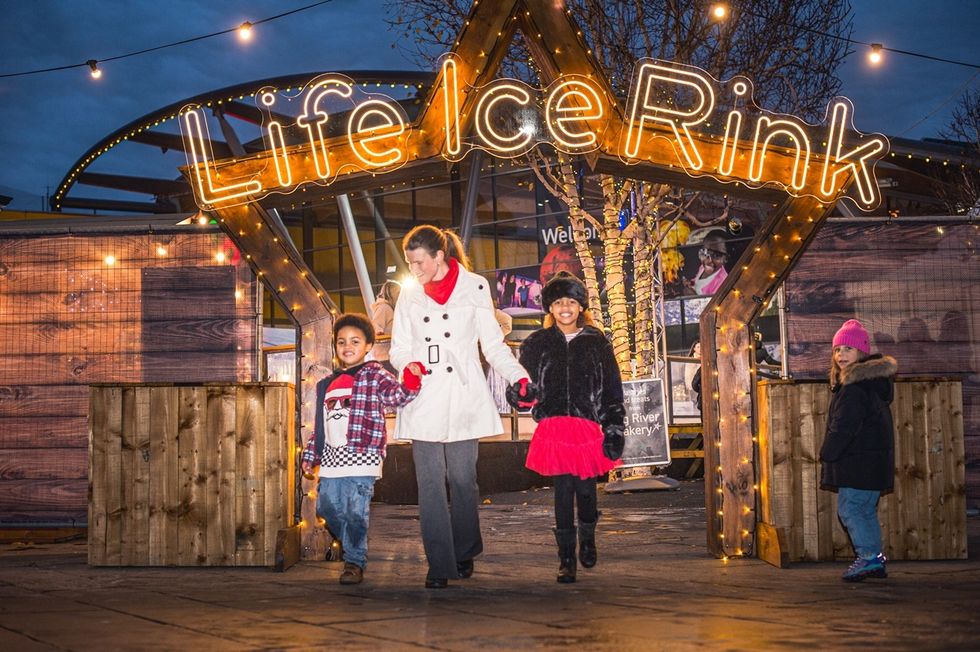 Family walking under a "Life Ice Rink" sign, surrounded by festive lights.