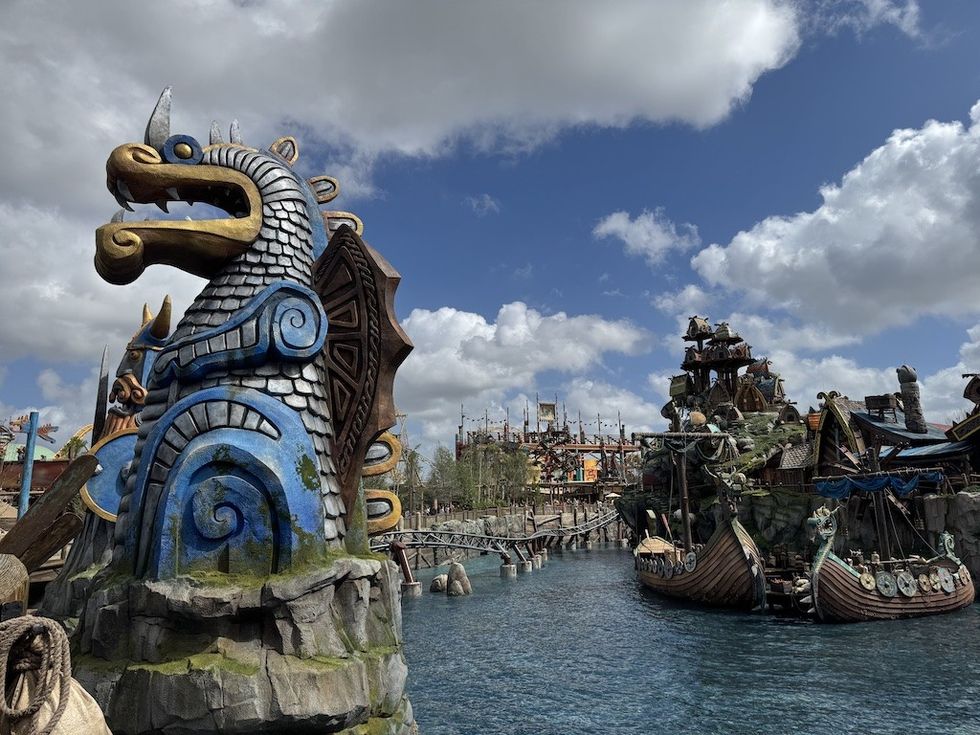 Fantasy dragon statue by a river, with cloudy sky and themed buildings in the background.