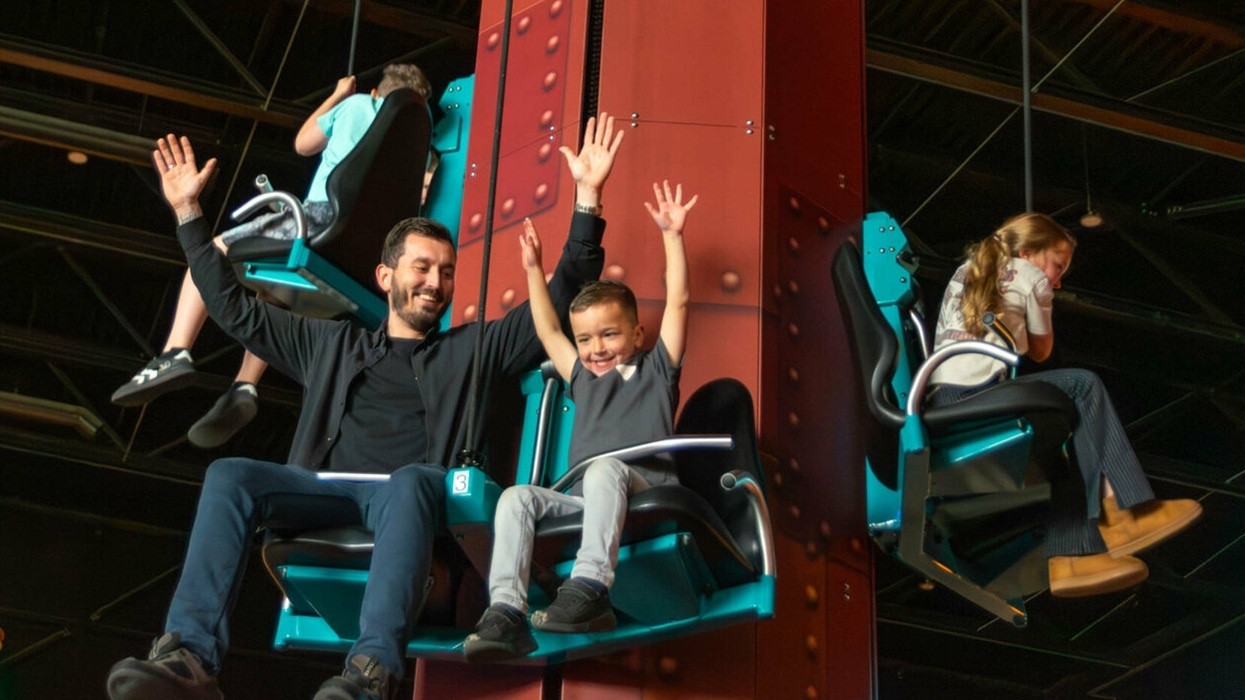 Father and son enjoy a drop tower ride at Bommelwereld, with hands raised; others ride beside them indoors.