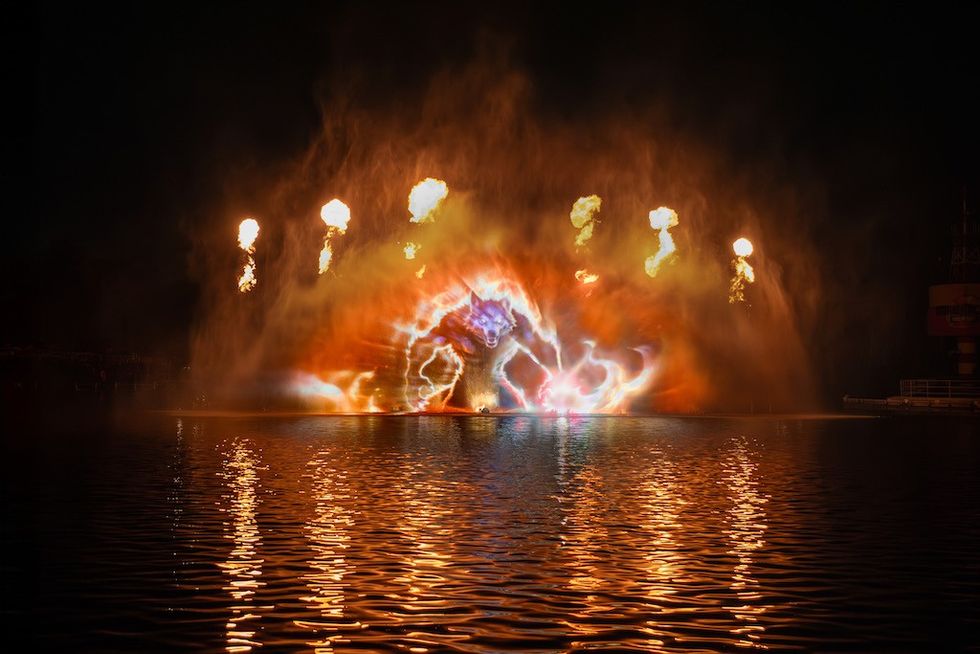 Fiery water show with colorful explosions and flame jets above a reflective lake surface.