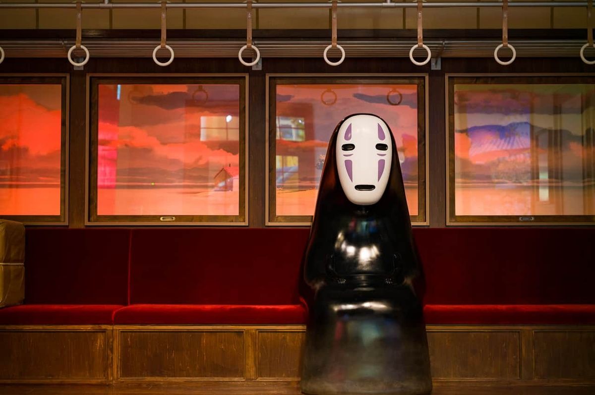Figure with white mask sits on red train seat against sunset backdrop.