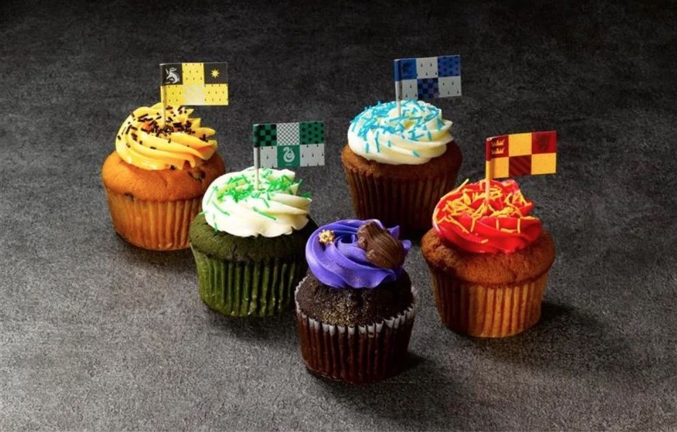 Five Hogwarts themed cupcakes with colored frosting and small flag toppers on a dark surface.