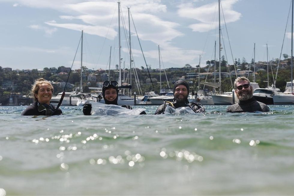 Four divers smiling in water near boats and a marina.