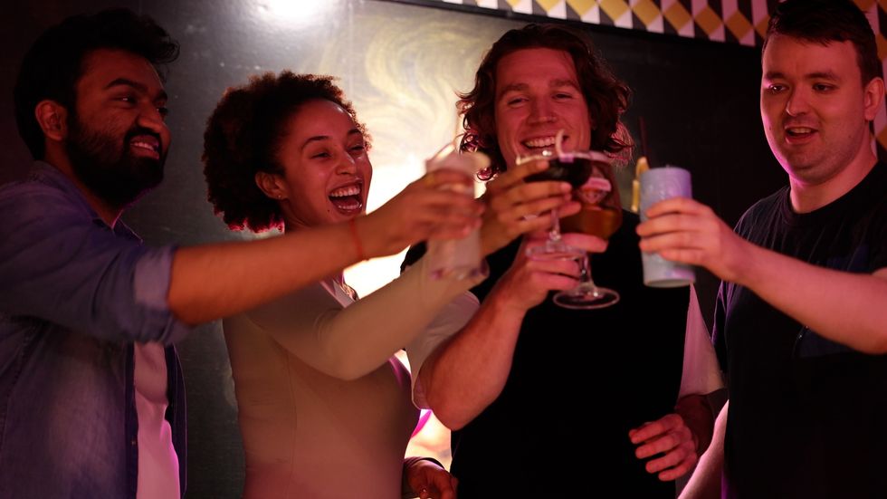 Four friends smiling and toasting with colorful drinks at a party.