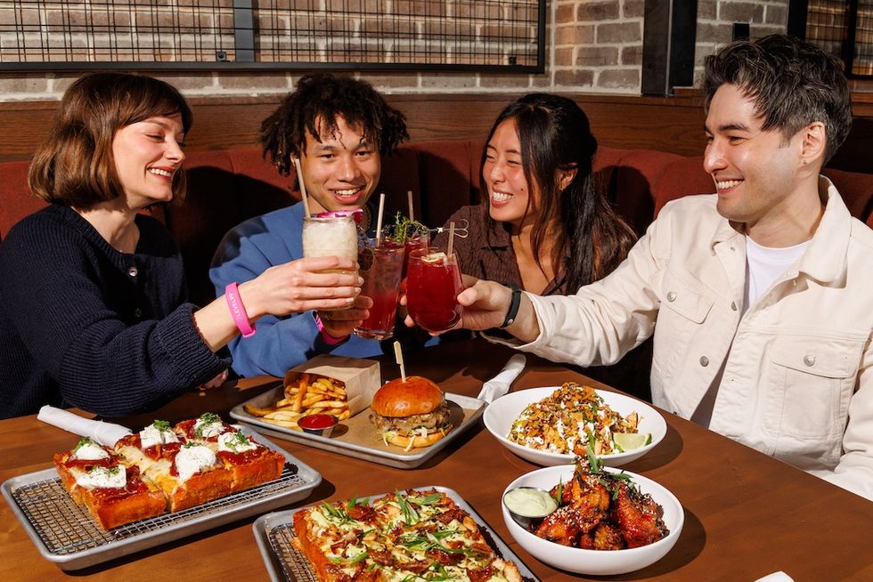 Four friends toasting drinks at a table with pizza, wings, and fries at Level99