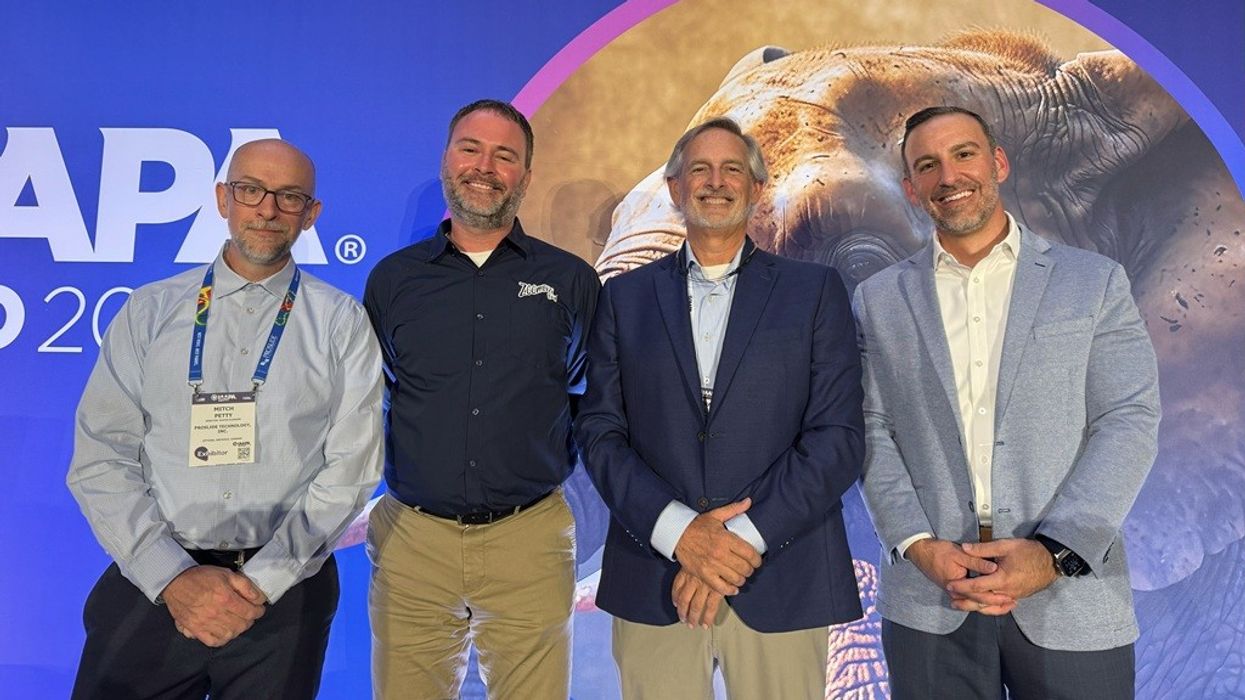 Four men in business attire standing together, smiling, with a conference backdrop.