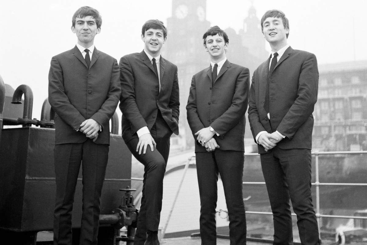 Four men in suits standing together on a dock in front of a historic building.