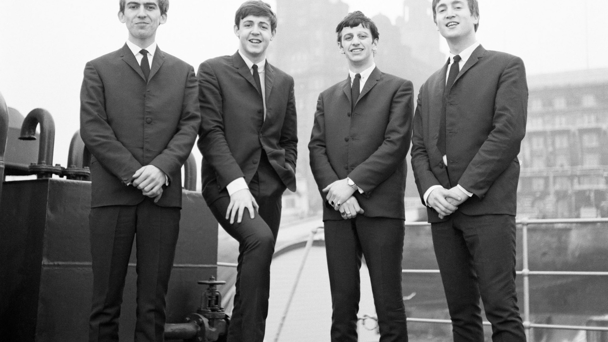 Four men in suits standing together on a dock in front of a historic building.