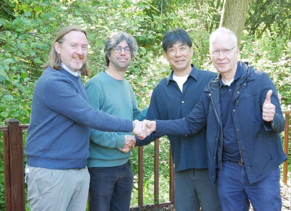 Four men smiling and shaking hands outdoors with a leafy background.
