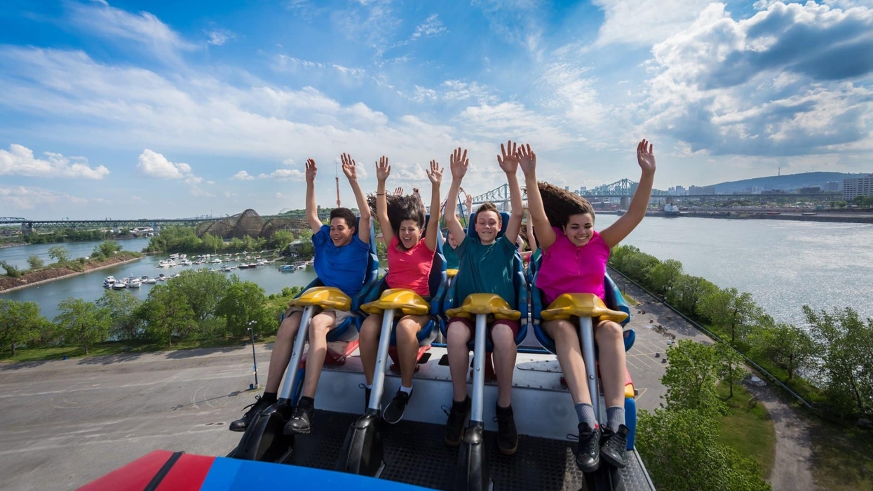 Four people enjoying a roller coaster ride with arms raised, clear sky and city in background.