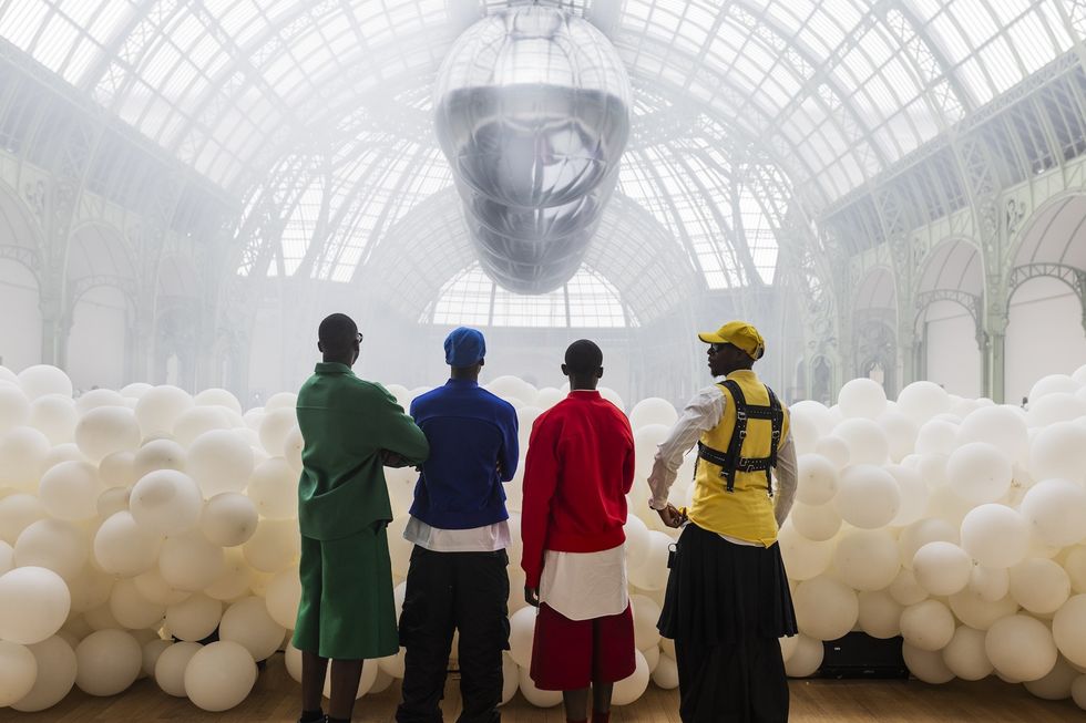 Four people in colorful outfits stand before white balloons and a large silver zeppelin indoors.