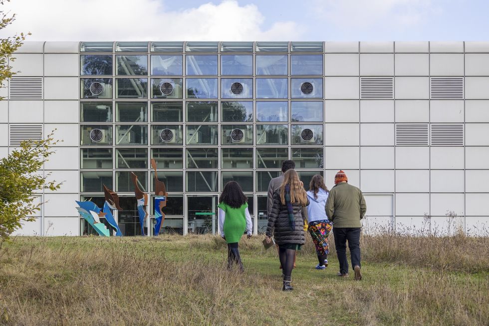 Four people walk towards a modern glass and metal building, the Sainsbury Centre in Norwich, after exploring the sculpture park