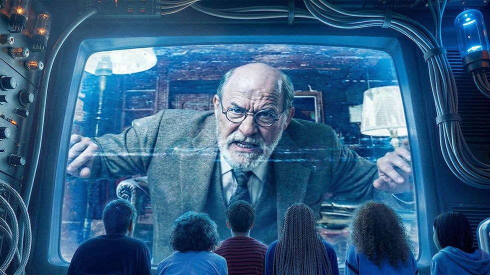 Four people watch a large screen displaying an older man speaking and gesturing.