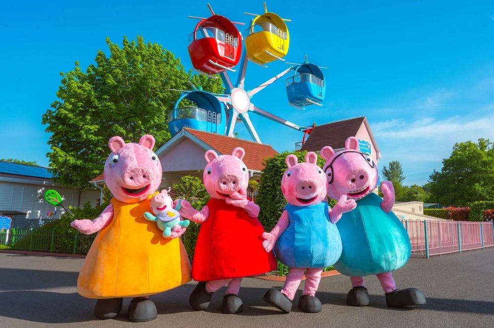 Four pig mascots in colorful outfits at a theme park with a Ferris wheel in the background.