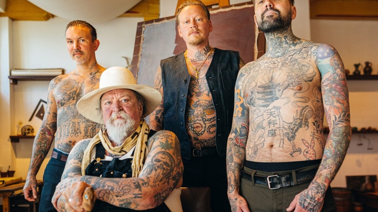 Four tattooed men, one seated in a hat; rustic interior background.