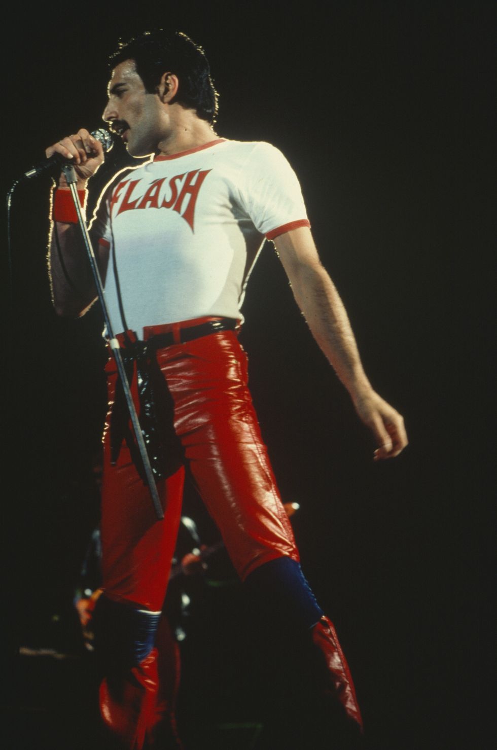 Freddie Mercury in a "Flash" shirt and red pants singing into a microphone on stage.