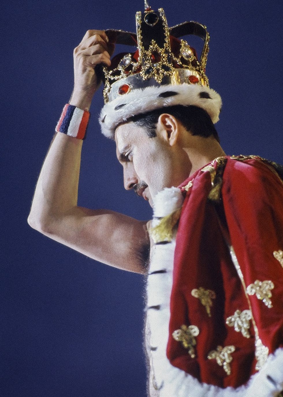 Freddie Mercury in red and gold cape wearing a crown, raising an arm with a tricolor wristband.
