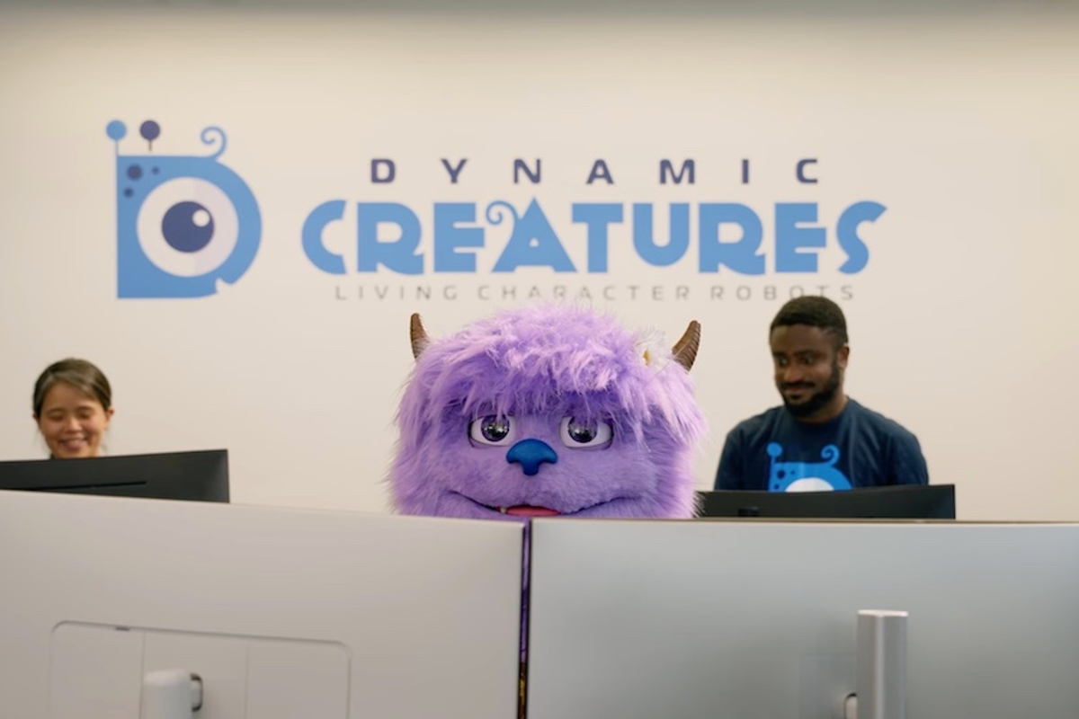 Furry purple creature at office desk with two people, under "Dynamic Creatures" sign.