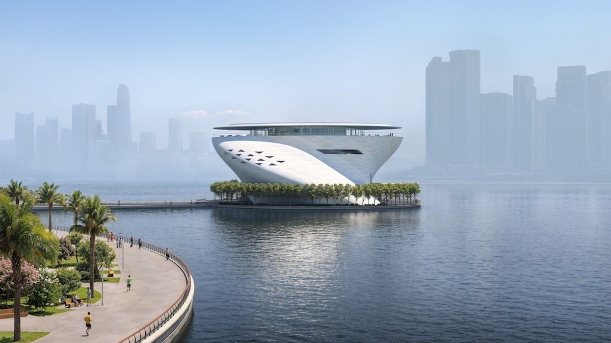 Futuristic building by waterfront surrounded by trees, with skyscrapers in the hazy background.