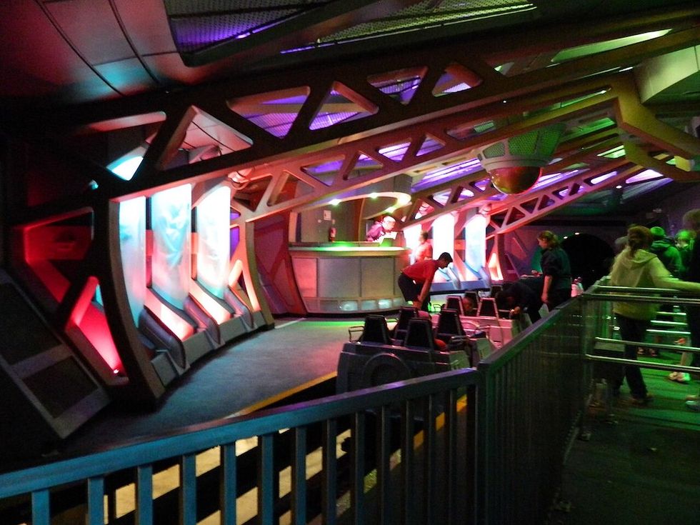 Futuristic indoor ride platform with colorful lighting and people boarding seats.