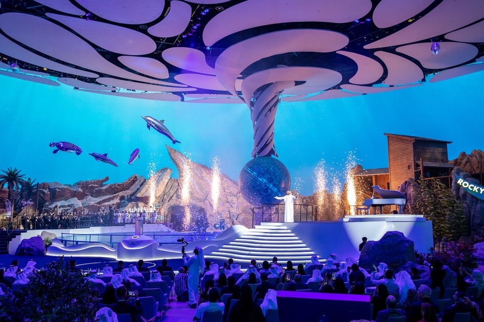 Futuristic stage with dolphins, swirling decor, and audience in an aquatic-themed performance.