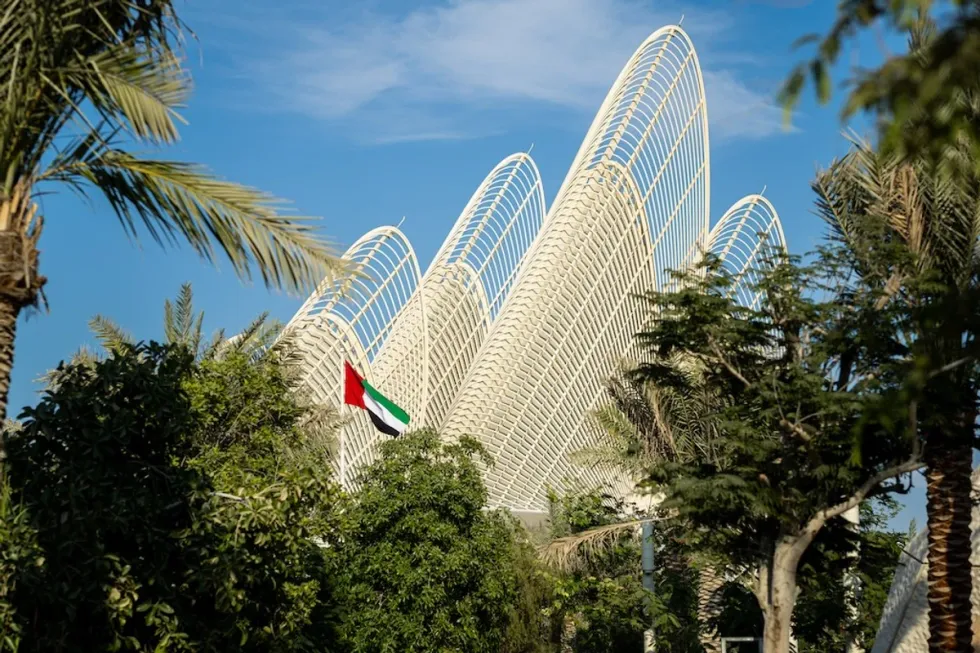 Futuristic white structure amid trees with a visible UAE flag; clear blue sky.