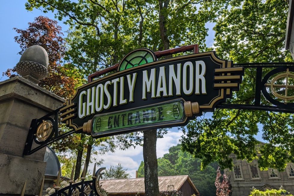"Ghostly Manor entrance sign with trees and old building in background."