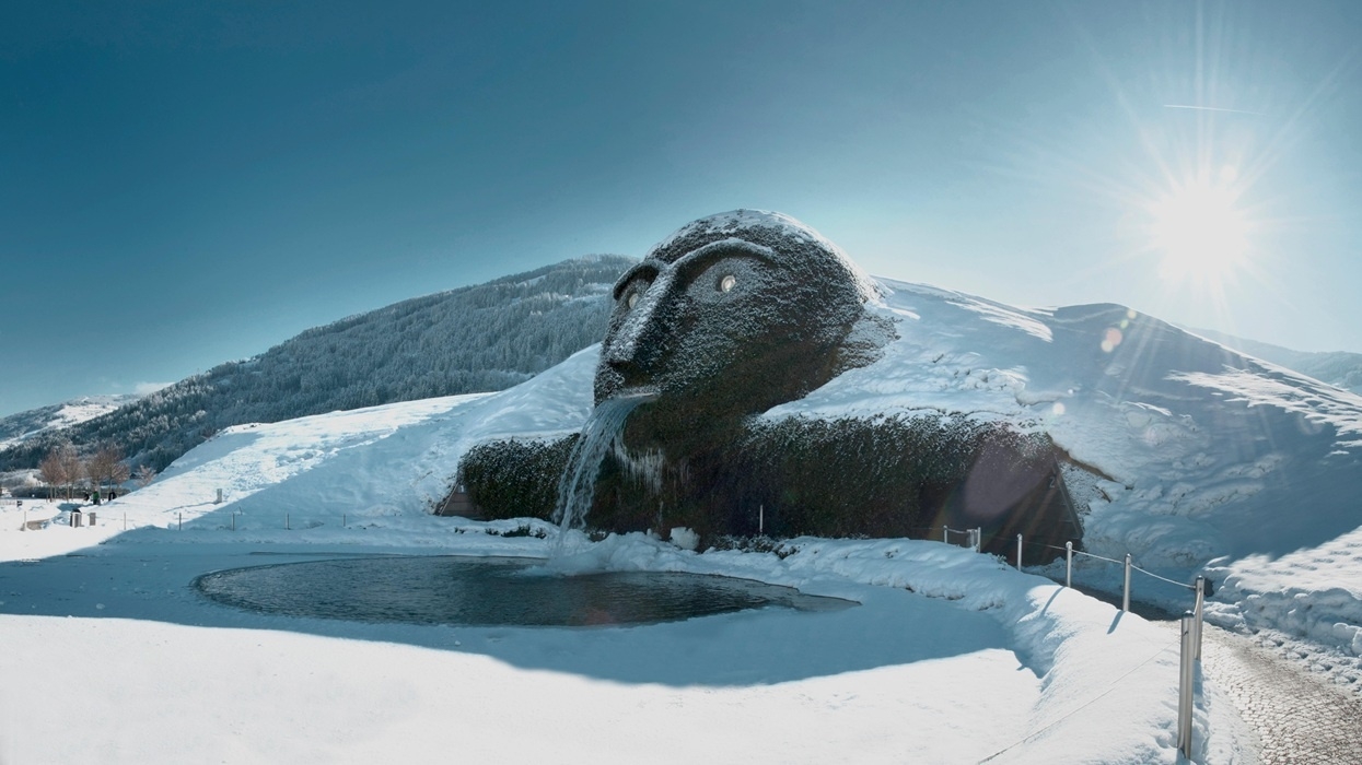 Giant head sculpture with waterfall mouth in snowy landscape under sun.