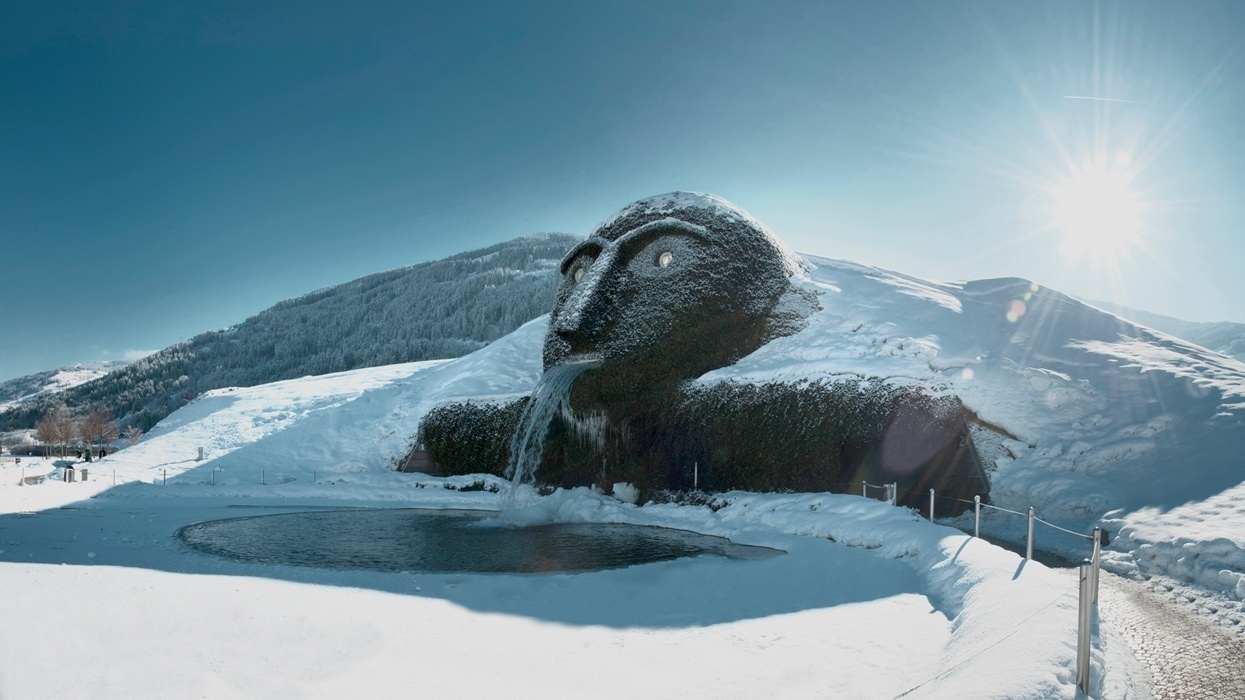 Giant head sculpture with waterfall mouth in snowy landscape under sun.