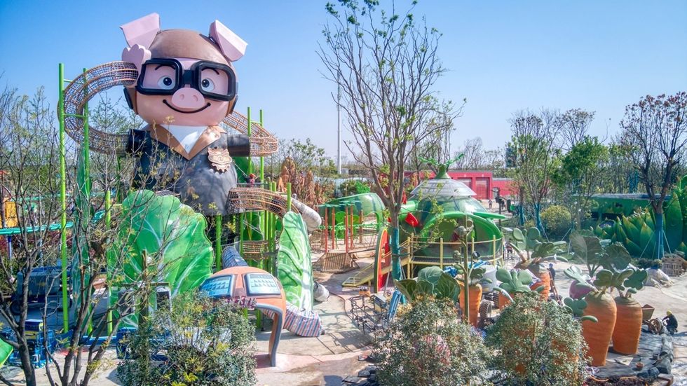 Giant pig sculpture with glasses in a whimsical outdoor park with plants and playground.