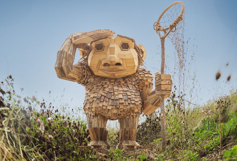 Giant wooden troll sculpture holding a net, standing in a grassy field against a blue sky.