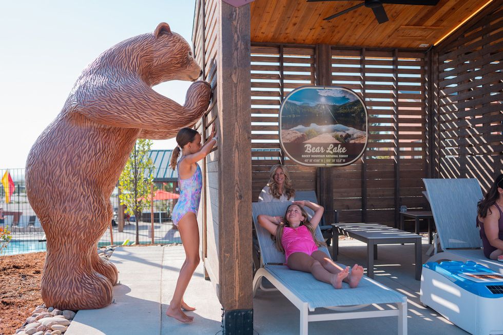 Girl beside bear statue, others relax on lounge chairs in shaded area near pool at Water World Summit Canyon