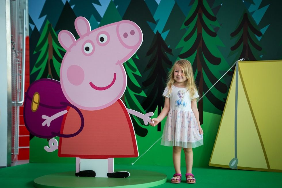 Girl holding hands with Peppa Pig cutout, standing in front of forest and tent backdrop.