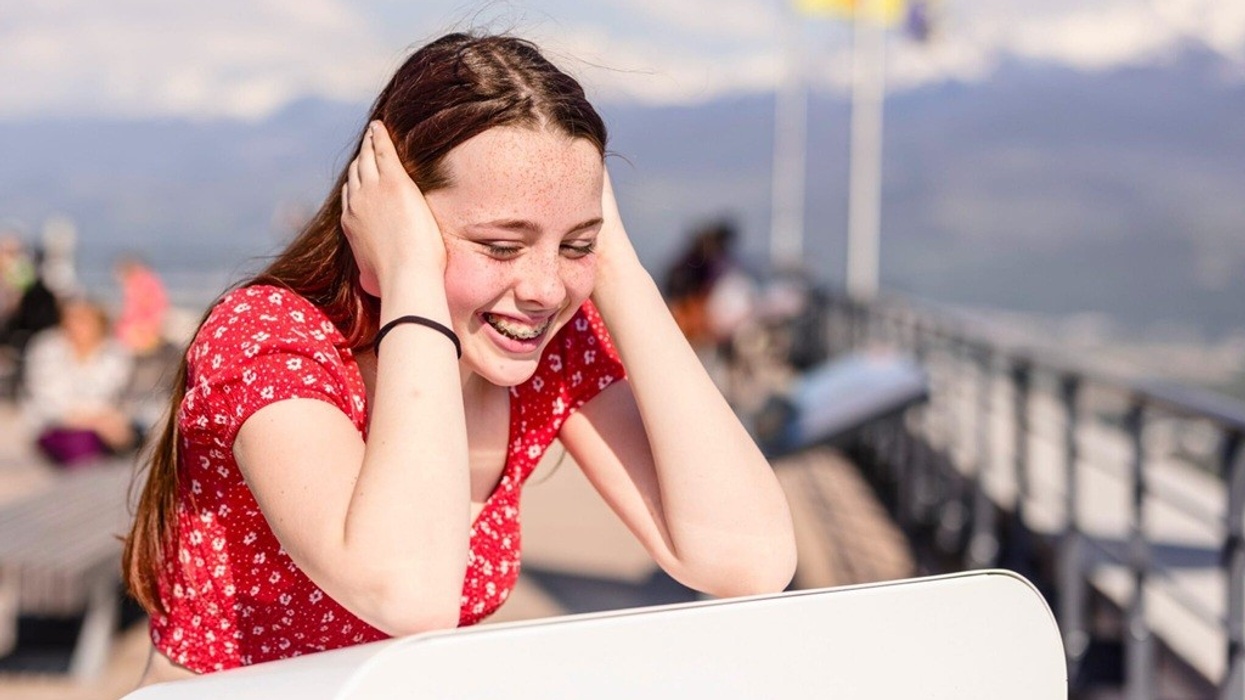 Girl in red shirt smiling, covering ears, outdoors with mountain view.