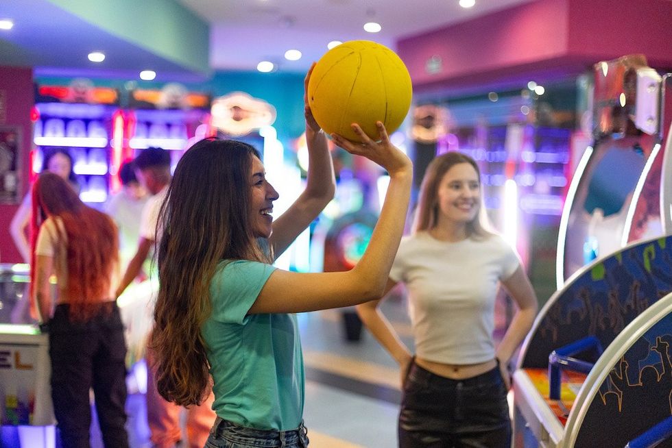 Girl playing basketball arcade game, friends watching, in a colorful game arcade.