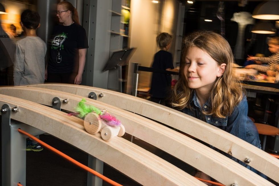 Girl watches a colorful wooden car roll down a track in a science exhibit.