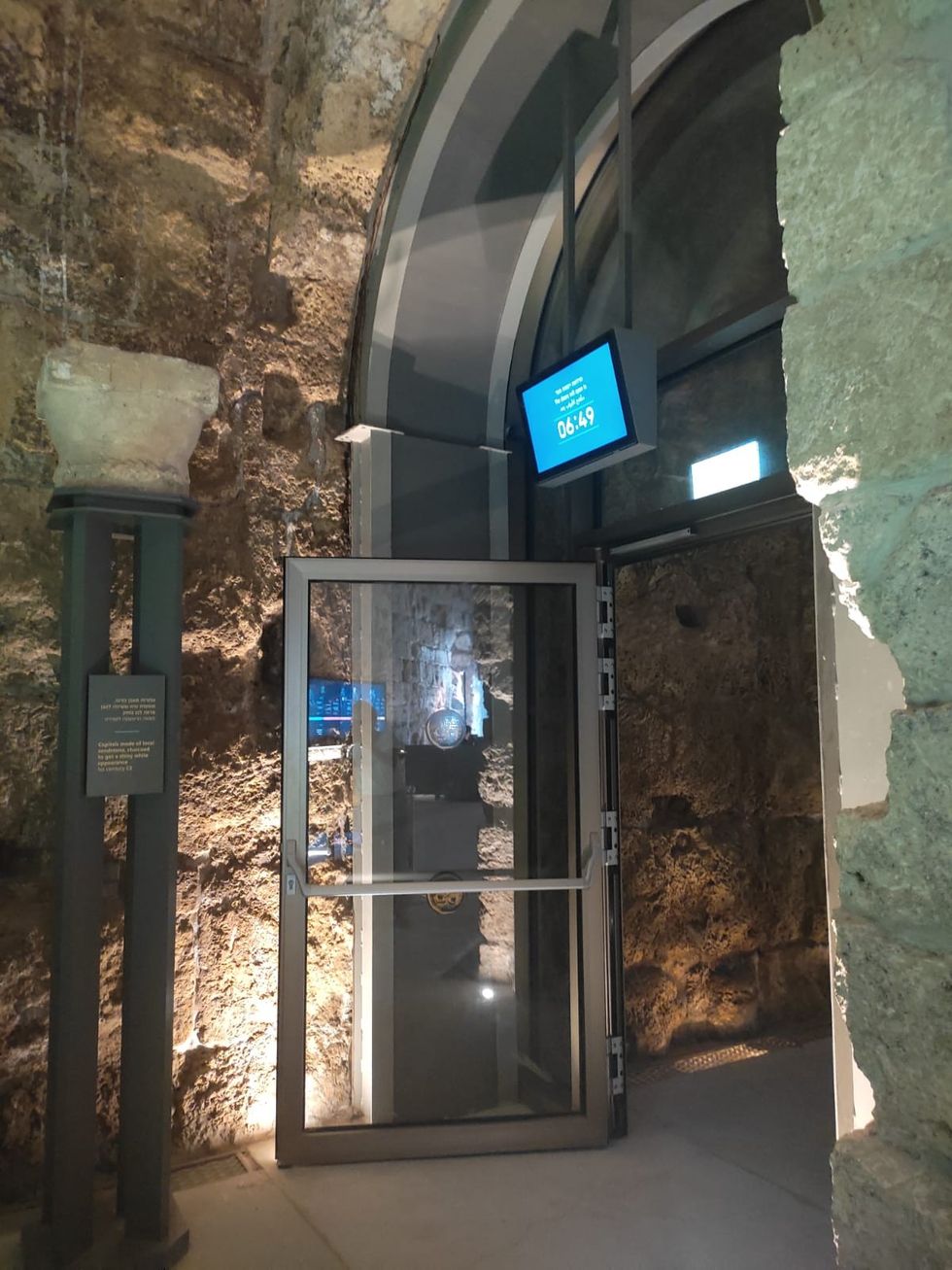 Glass door in ancient stone corridor, digital clock reads 06:49 at Caesarea Harbour Visitor Centre