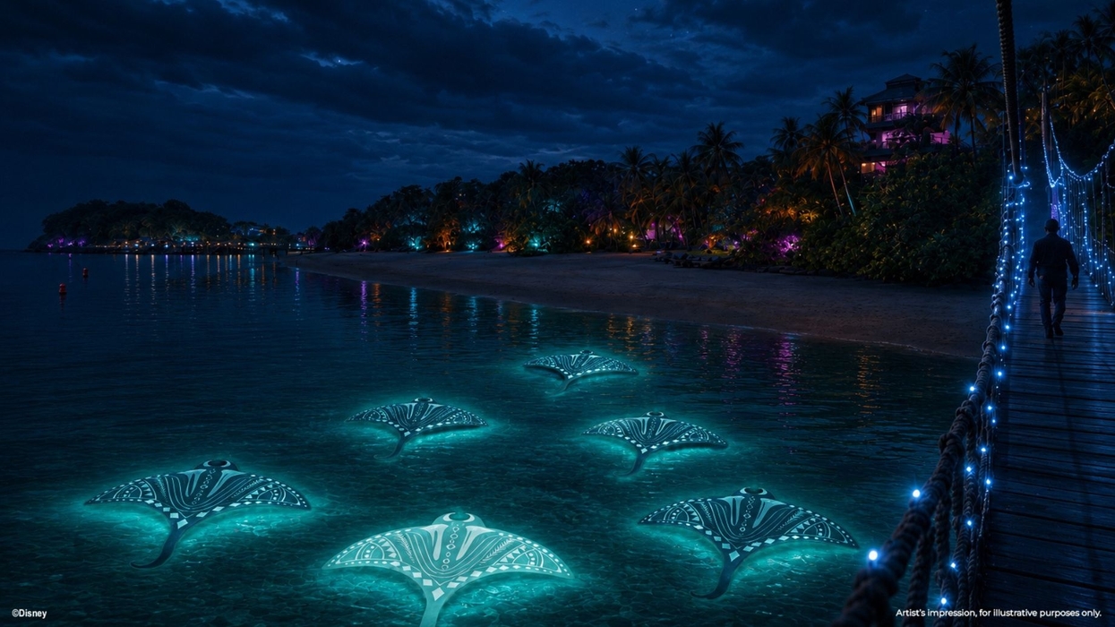 Glowing manta rays swim near a lit bridge and colorful shoreline at night.