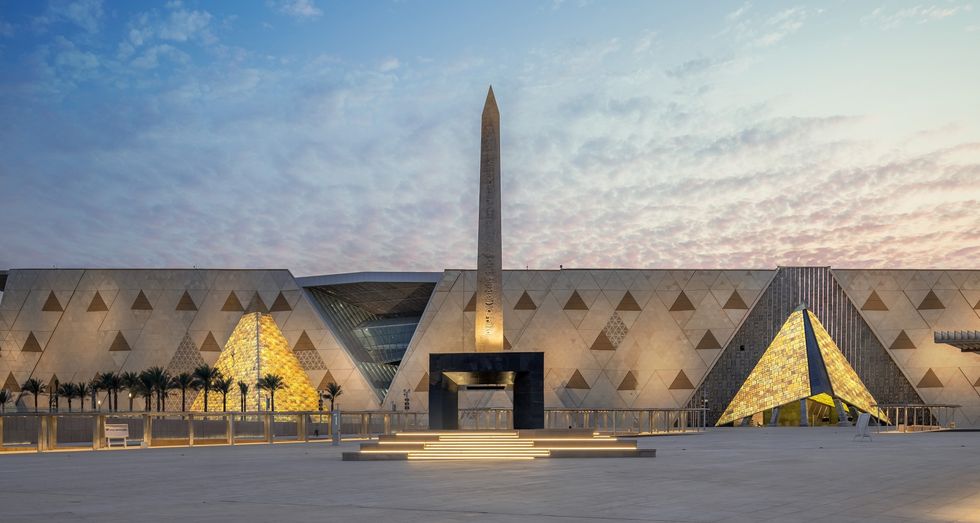 Grand Egyptian Museum facade with an obelisk and glowing pyramid structures at sunset.