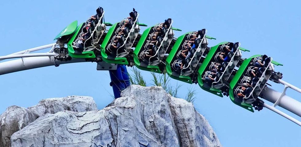 Green roller coaster with passengers soaring over rocky terrain against a clear blue sky.