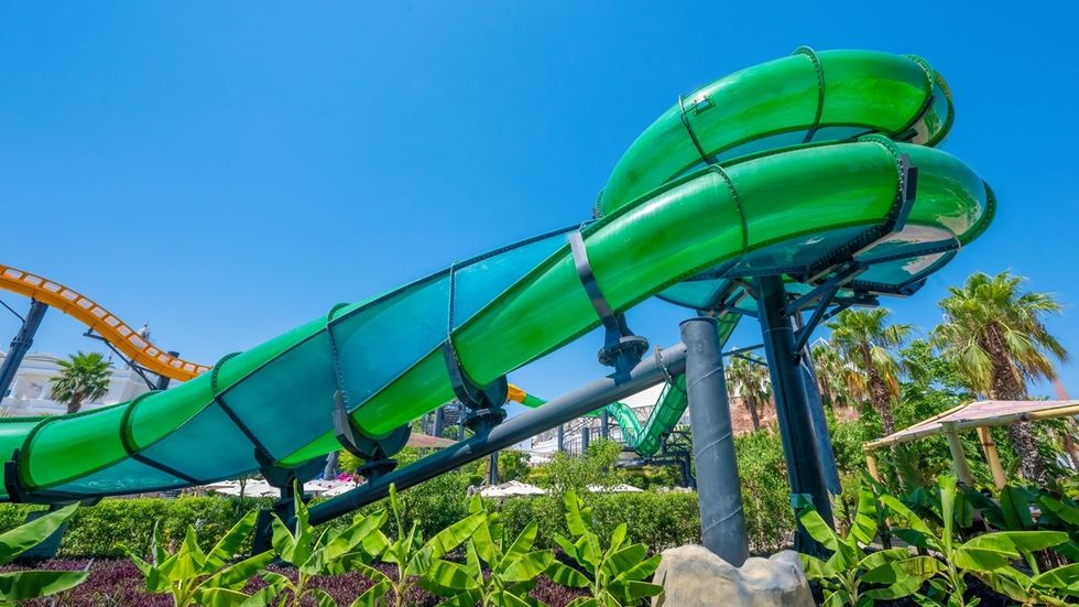 Green water slide with a Helix Twist curving in a sunny amusement park named The Land of Legends, surrounded by tropical plants.