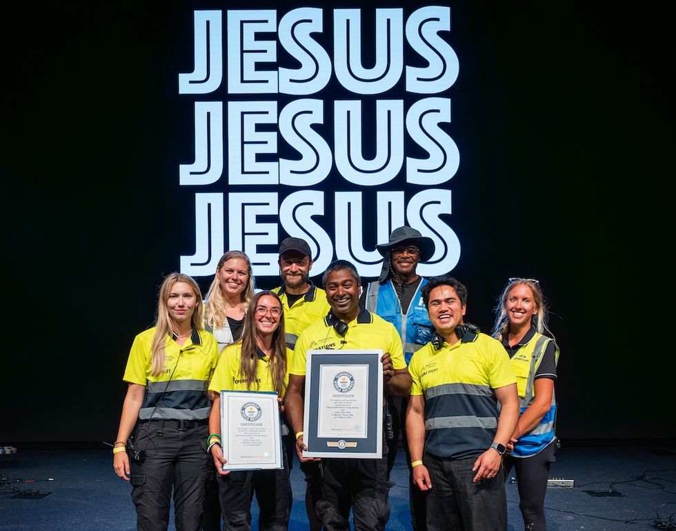 Group from Aerial Illuminations in yellow shirts holds Guiness World Records certificates, stands before "JESUS" sign.
