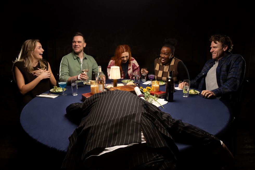 Group laughing at a table while a person in a pinstripe suit lies face down in front.